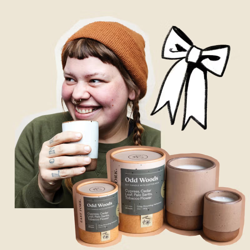 A person with a nose ring and tattoos on their fingers, wearing an orange beanie, smiles while holding a white cup. In front of them are various sizes of Odd Woods candles with labels, and a black-and-white illustration of a ribbon is in the background.