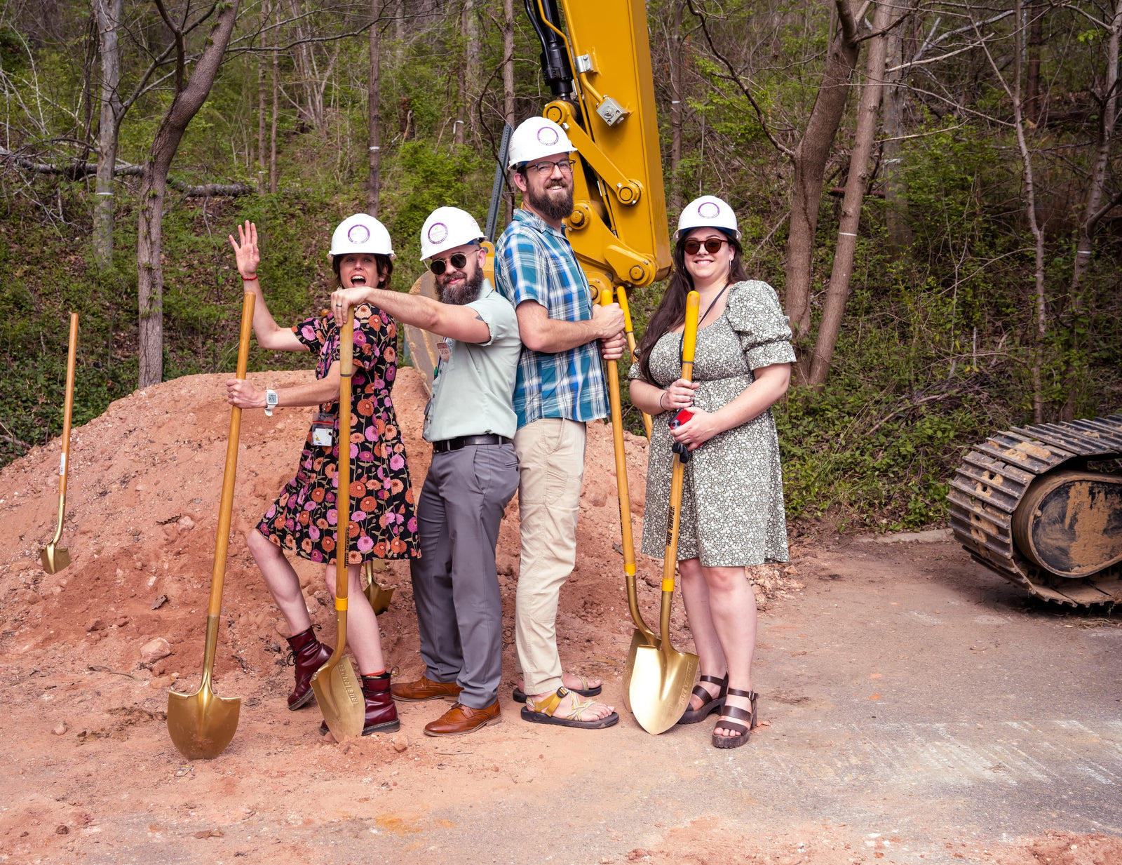 Four people with shovels and hard hats in front of an excavator, likely at a groundbreaking ceremony.