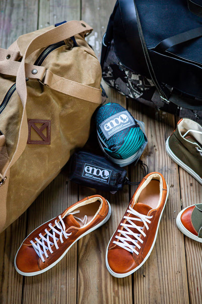 A wooden floor is scattered with travel essentials, including a tan canvas duffel bag, a pair of brown leather sneakers with white laces, and ENO DoubleNest Hammock and Atlas Suspension System bags.