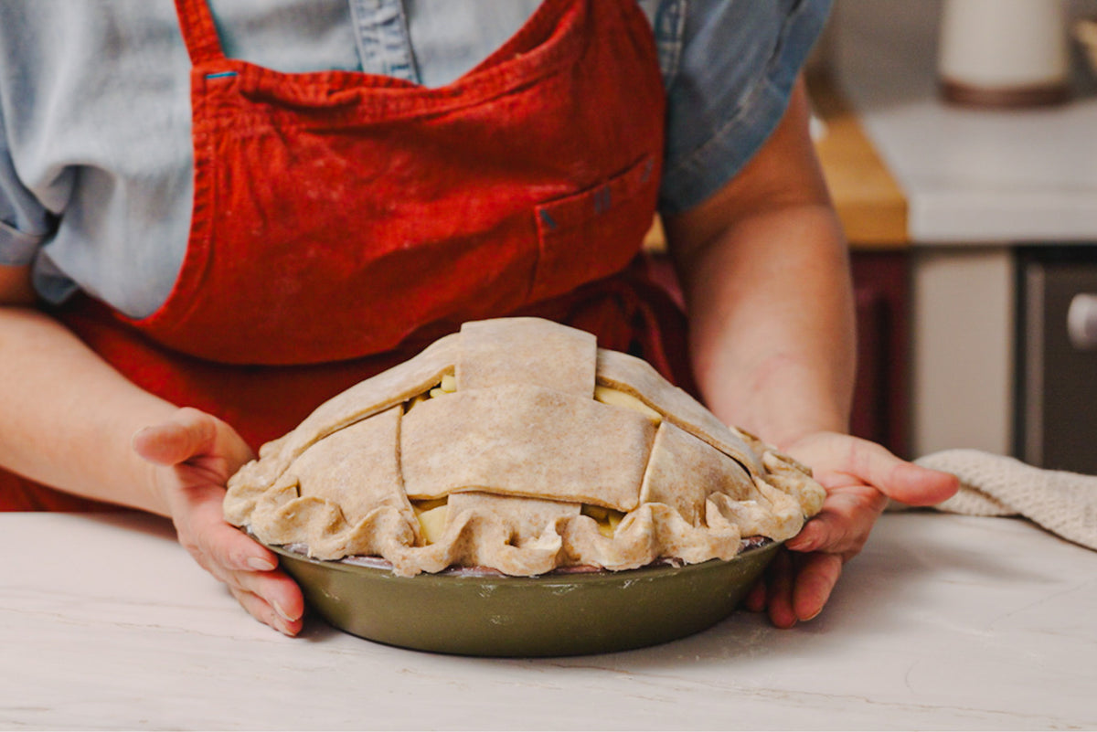An uncooked pie with a lattice crust and crimped edges sits in a ceramic green pie dish on a kitchen counter, held by a person wearing a red apron.