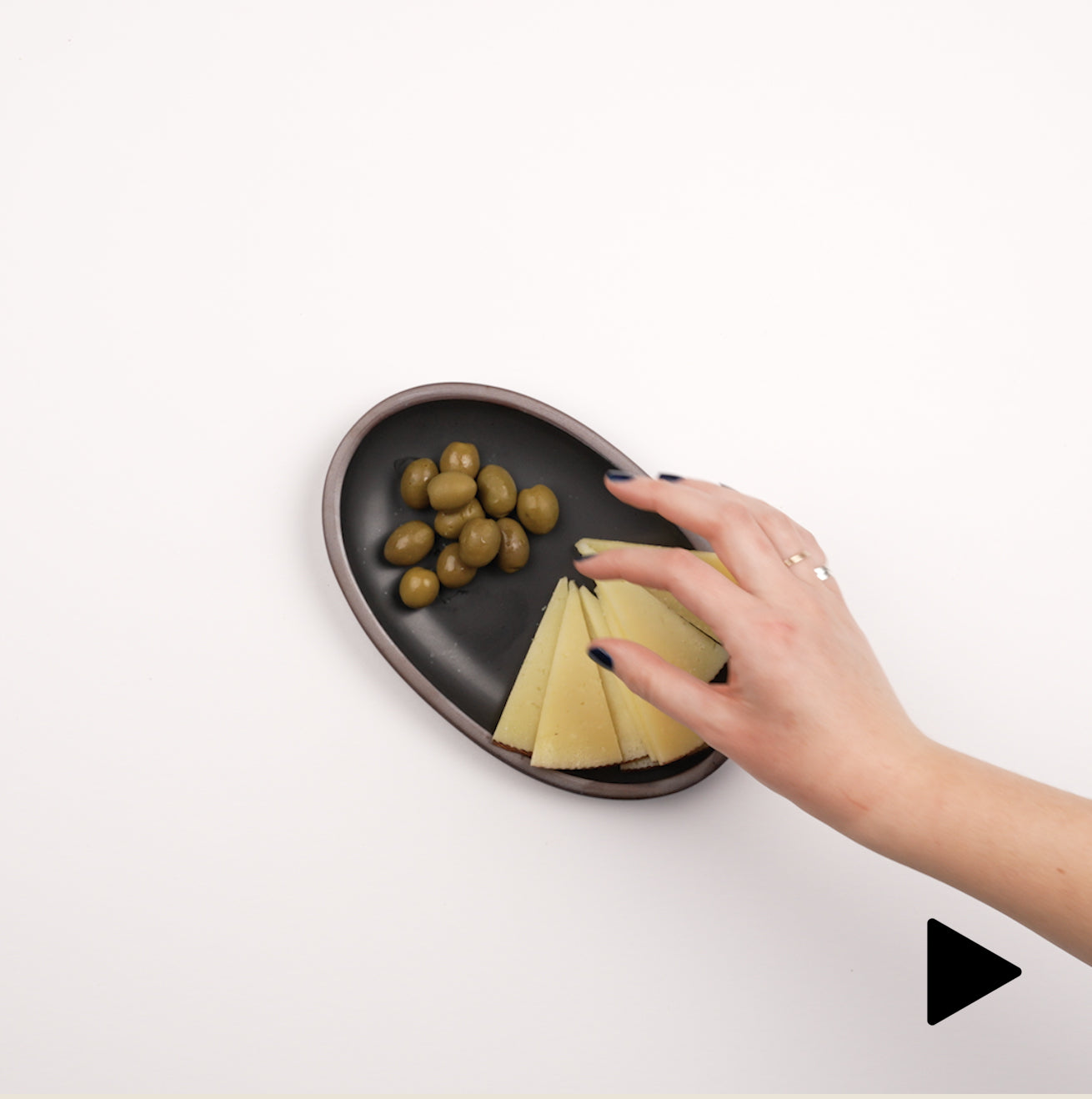 Video of a hand placing a small oval platter in a blue color down filled with olives and cheese slices, and a hand grabbing an olive.