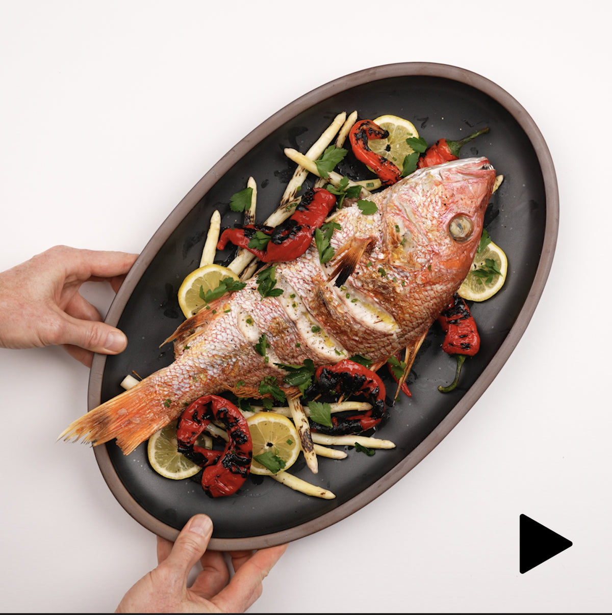 Video of a hand placing a large oval platter in a black color down with a fish entree, and a hand sprinkling salt.