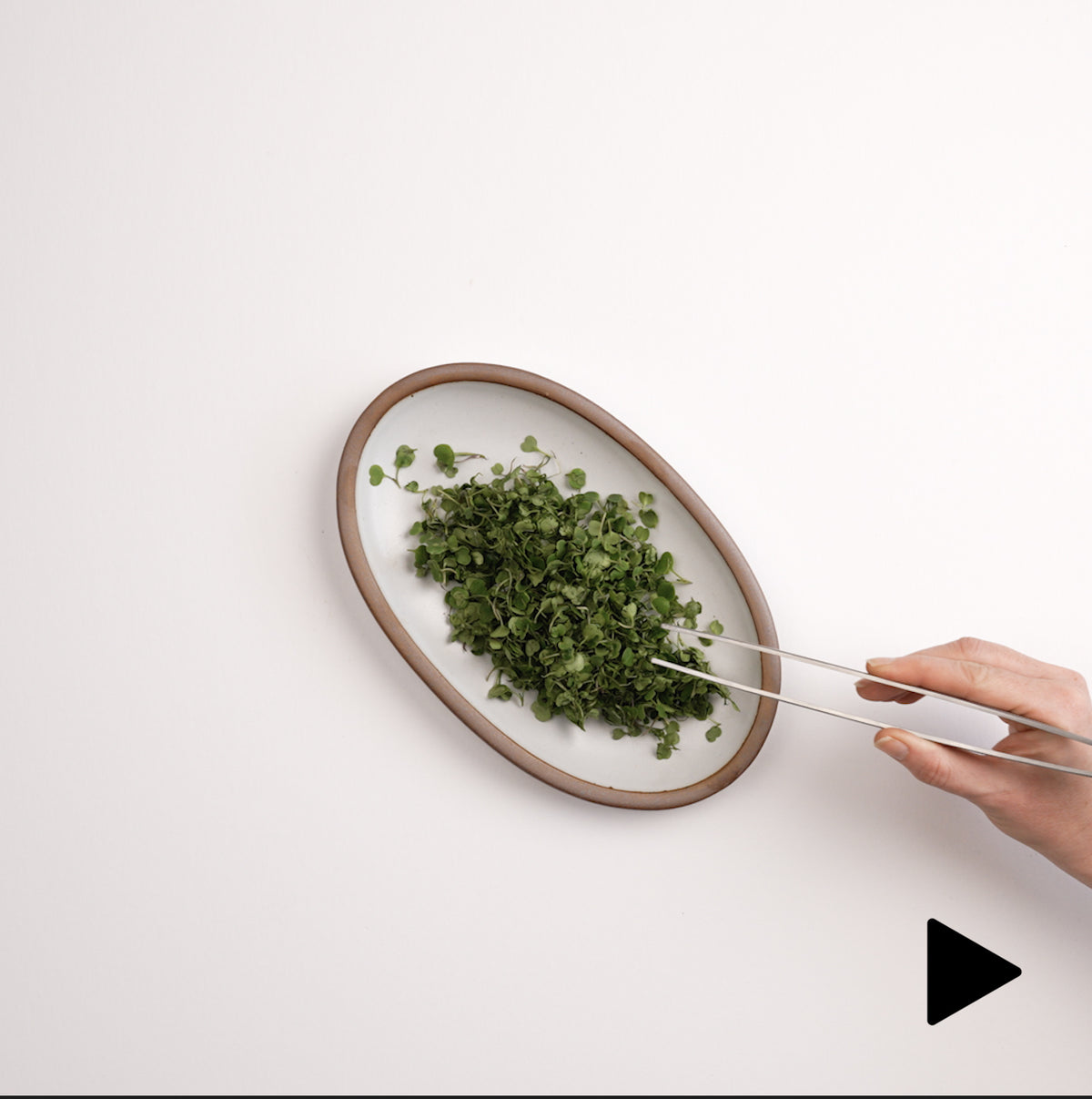 Video of a hand placing a small oval platter in a white color down filled with chopped herbs, and a hand grabbing some with tiny tongs.