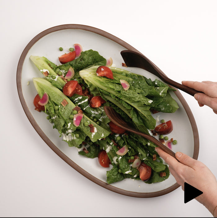 Video of a hand placing a large oval platter in a white color down with salad, and a hand using wood utensils to grab a serving.