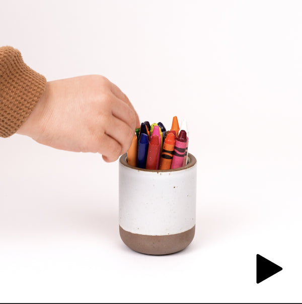 Video of a hand placing a tiny cup in a white color filled with crayons, with a child's hand grabbing a crayon.