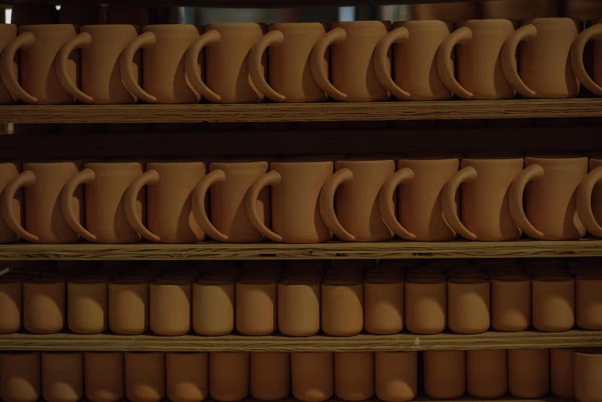 Rows of unglazed clay mugs are neatly arranged on shelves, showcasing the pottery process.
