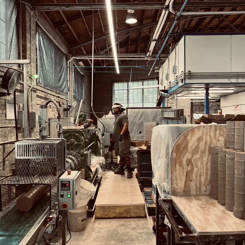 A worker stands in a ceramics studio using industrial equipment to process clay. The space features exposed brick walls, overhead lighting, and various tools and materials arranged throughout.