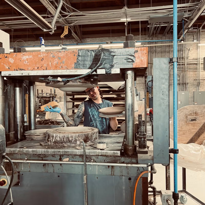 A person wearing a tank top and gloves operates a ram press in a ceramics workshop, holding a ceramic piece. The background features shelves and industrial equipment.