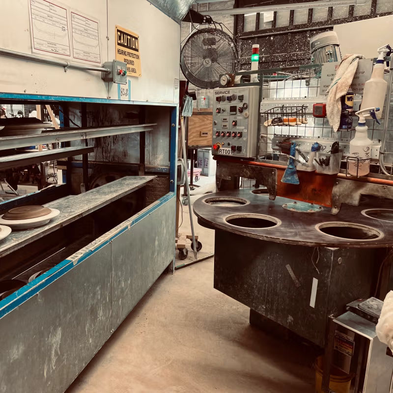 Industrial workspace with pottery equipment and tools, including a shelving unit with stacked plates and a control panel with various levers and buttons. The area is equipped with a large fan and labeled signs, emphasizing safety.