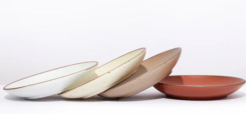 A row of five ceramic plates leaning against one another, featuring a variety of muted glazes including white, cream, beige, and terracotta, set against a plain white background.