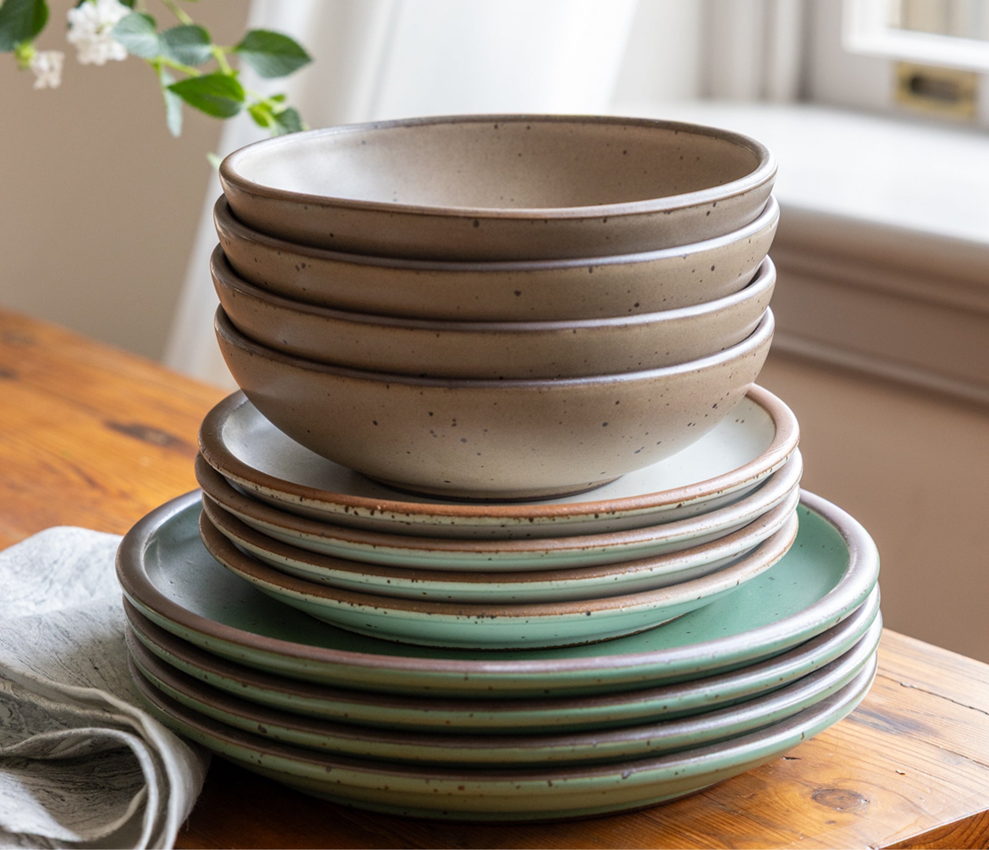 Stacked speckled ceramic bowls and plates in earthy green and taupe glazes on a wooden table.