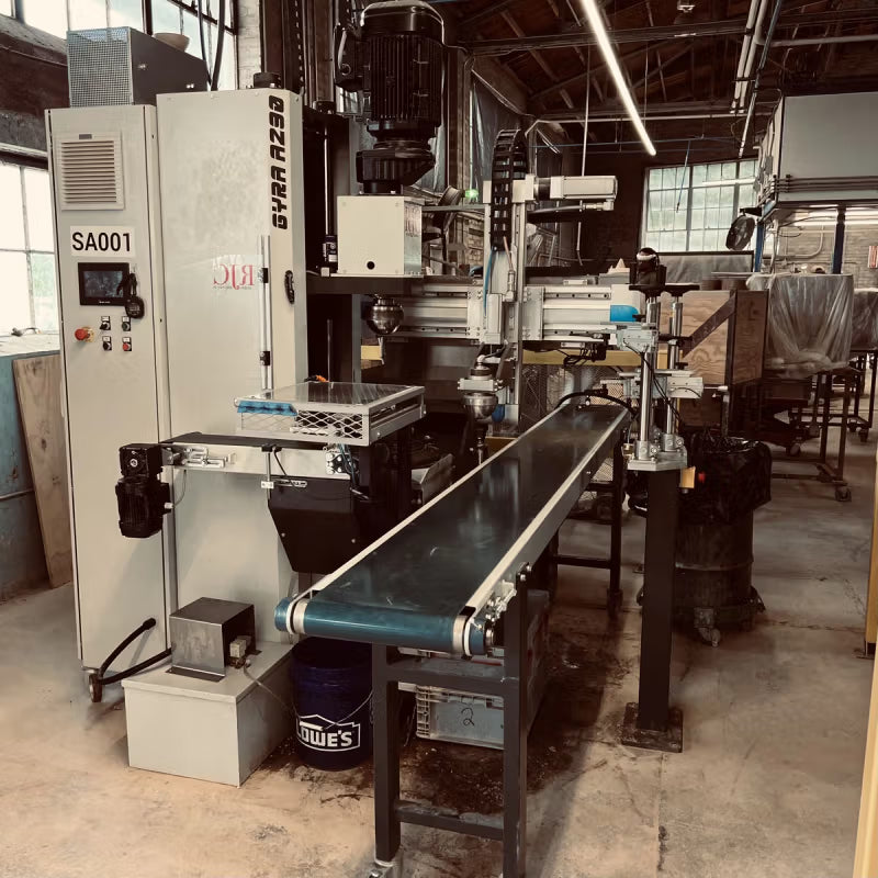 Industrial machinery and conveyor belts in a workshop setting, surrounded by large windows and overhead lighting.