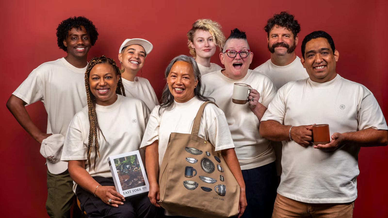 A diverse group of eight people stand together in front of a red background, all smiling. They wear matching light-colored shirts, and some hold East Fork ceramic mugs and a tote bag.