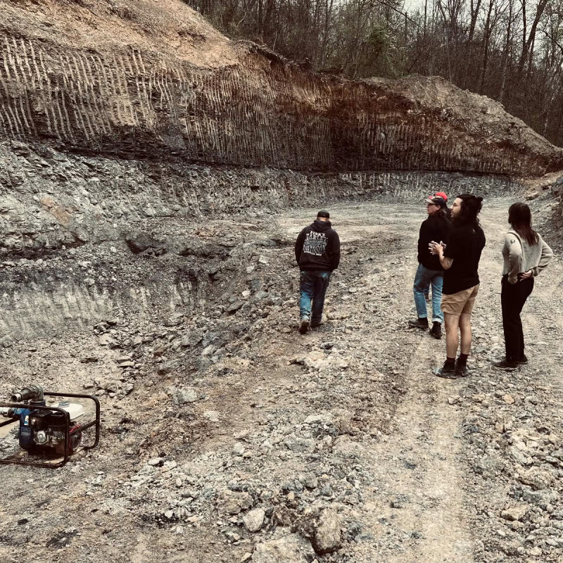A group of people stand in a rocky excavation site, with layered earth visible on the sides and a generator on the ground, surrounded by bare trees in the background.