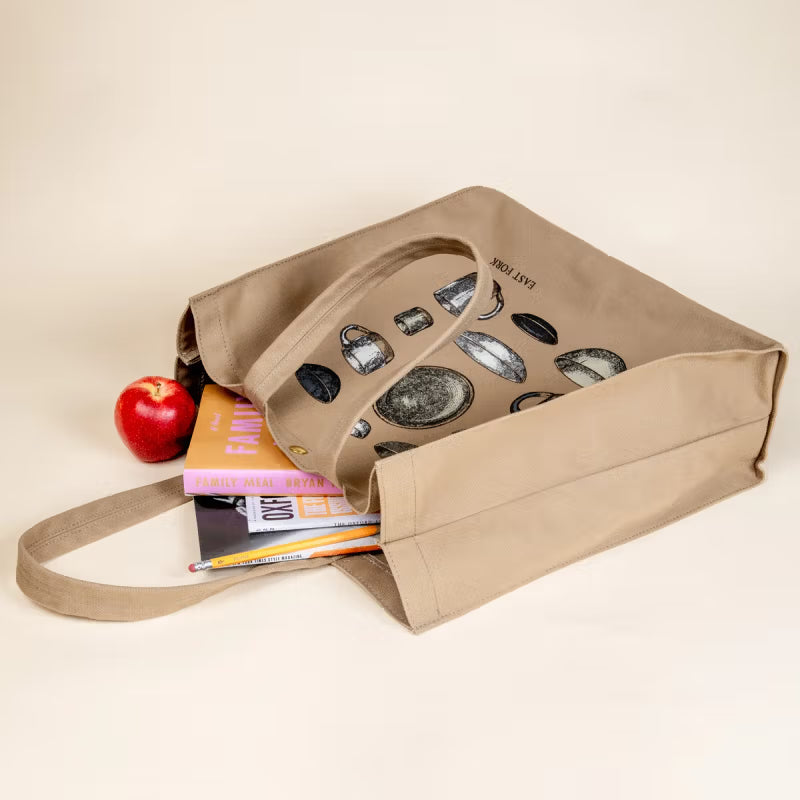 A beige tote bag featuring illustrations of pottery, partially filled with books and pencils, lies on its side next to a red apple on a light background.