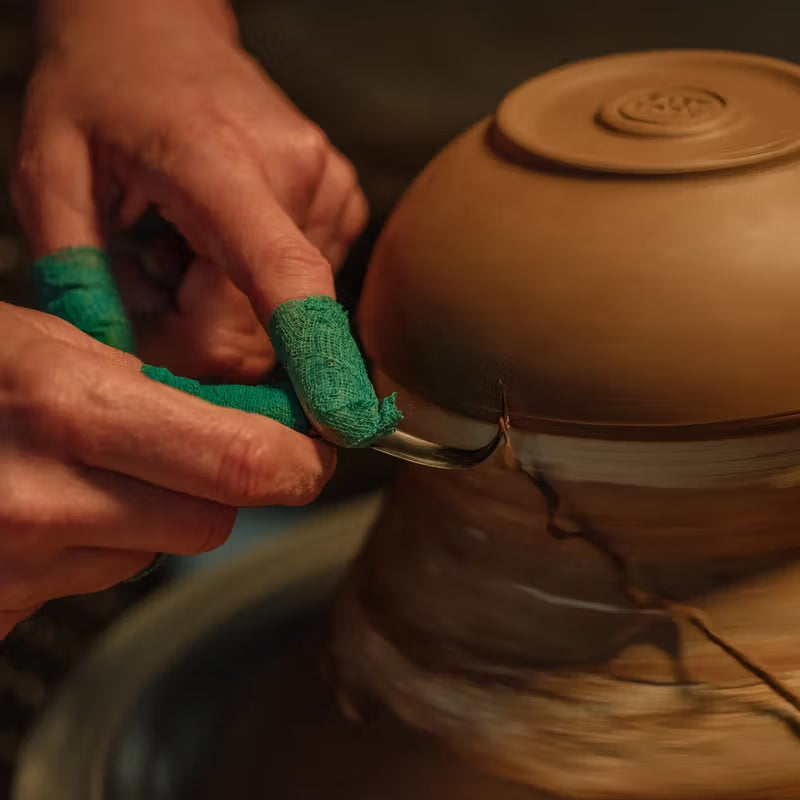 Hands with green protective wraps are using a trimming tool on the base of a clay pottery piece, focusing on refining its shape on a pottery wheel.