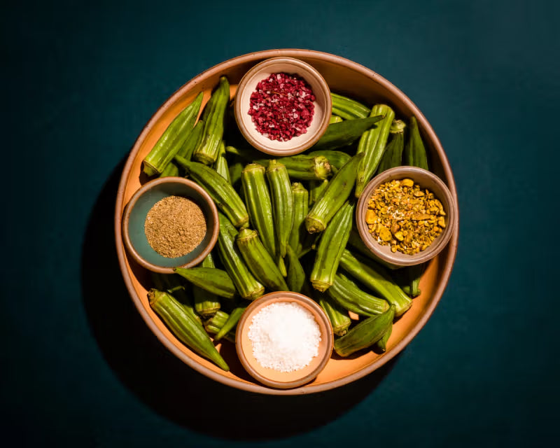 A terracotta bowl filled with fresh okra, accompanied by small bowls containing various spices and salts, set against a dark background.