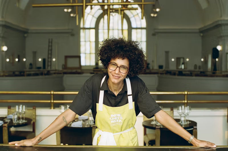 A person wearing glasses and an apron stands confidently leaning on a railing in an elegantly designed interior with large arched windows and a high ceiling.