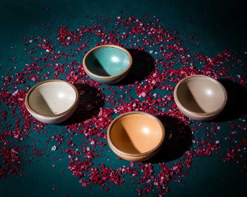 Four small ceramic bowls in various earth tones and a turquoise are arranged on a teal surface, surrounded by scattered pink salt crystals.