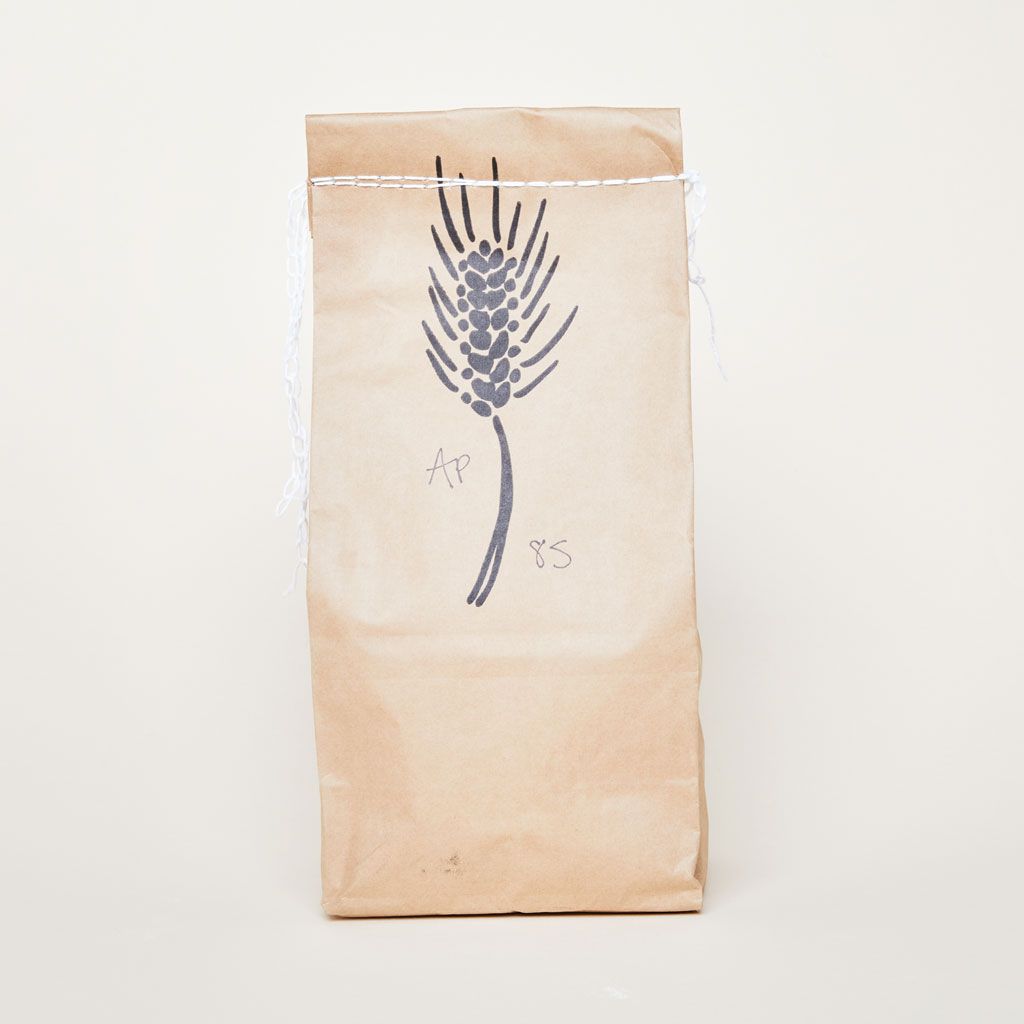 A brown paper bag stitched closed with white string at the top, a drawing of a piece of wheat and "AP 85" written on it