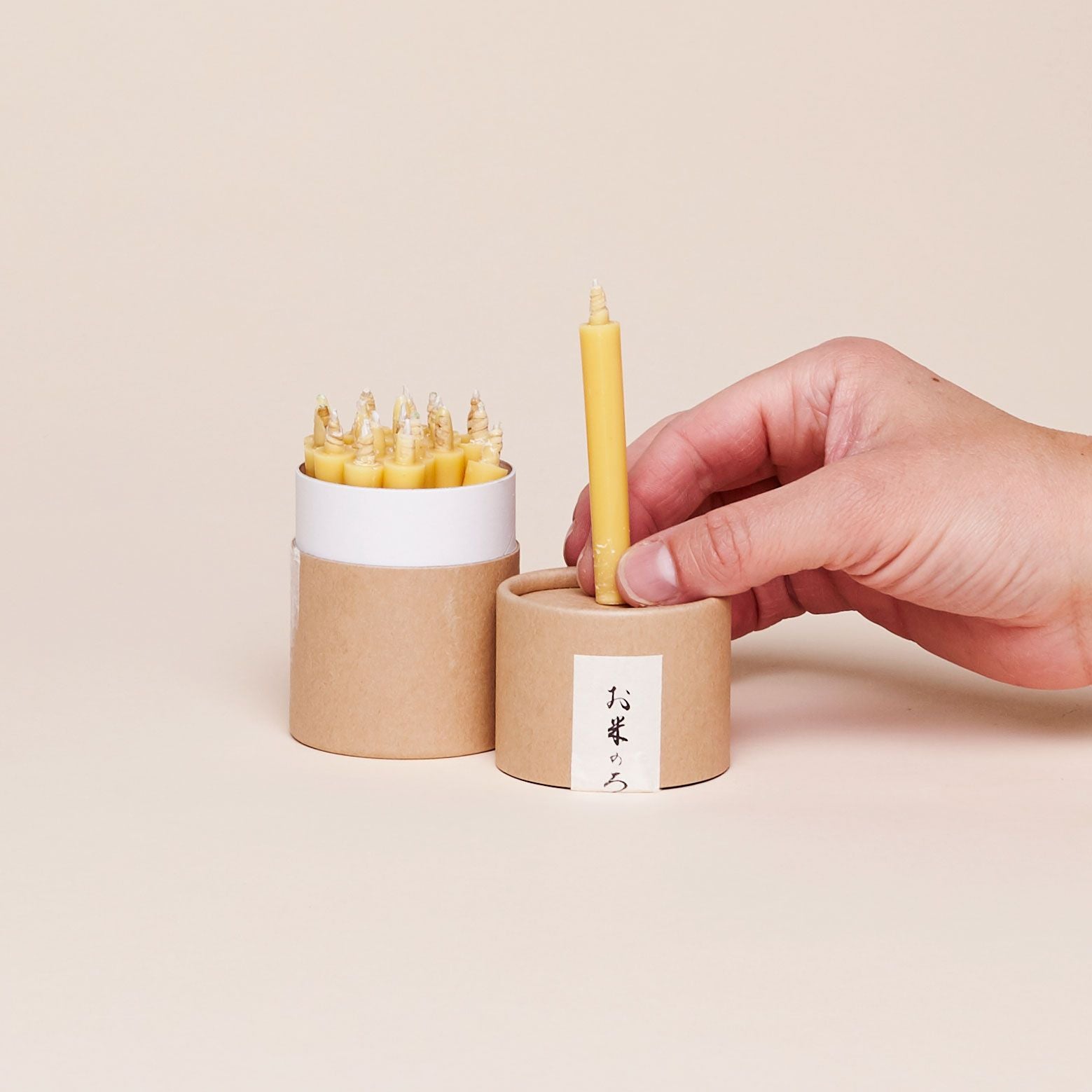 A brown and white cylinder that is open at the top holding yellow candles next to a hand with two fingers gripping the base of a candle that is identical to the others, atop a shorter brown cylinder