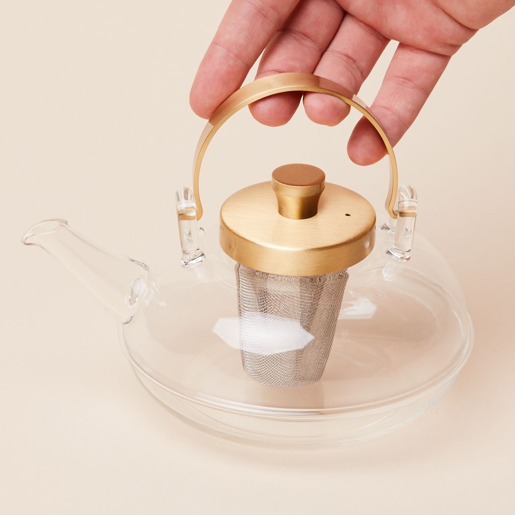 Fingers touch the gold handle of a tea pot made of glass with a glass spout that also has a gold lid and a gray metal silver insert for loose tea